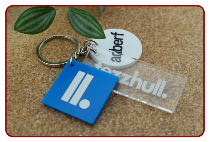 Blue square keychain with white logo and black dot, made of plastic.
